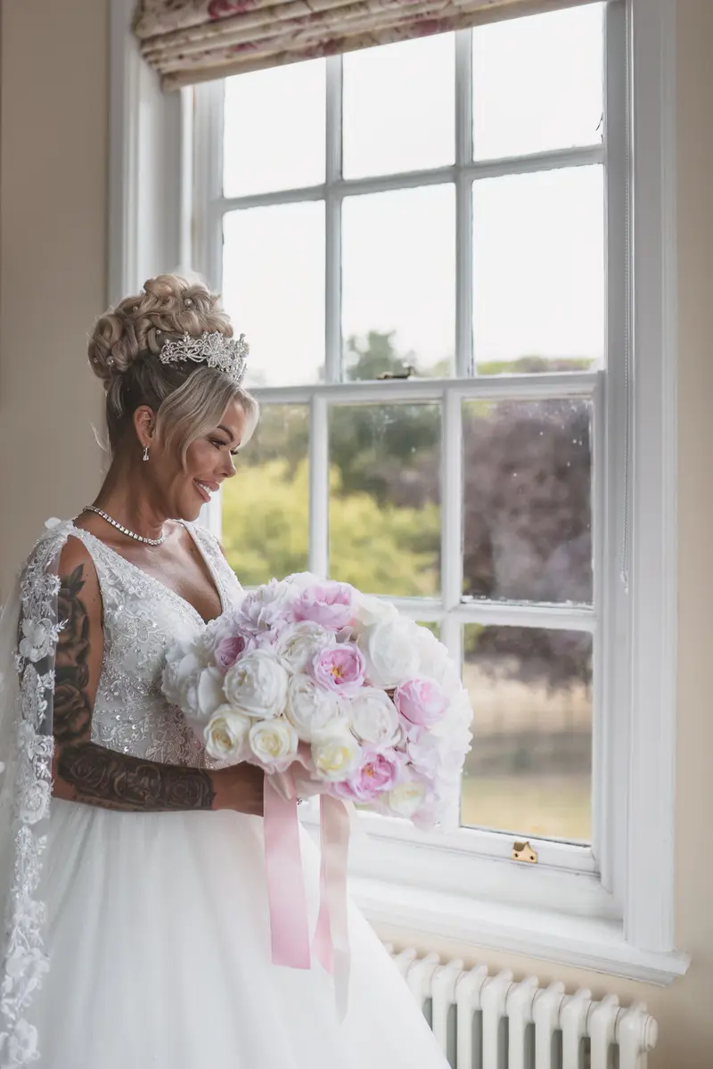Bride smiling with bouquet by window, romantic bridal morning photography, luxury UK wedding