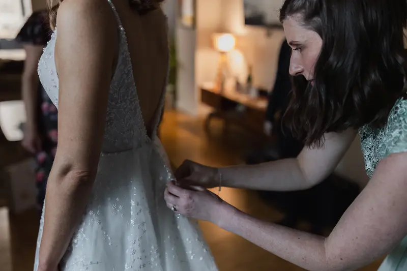 Bridesmaid helping bride fasten elegant wedding dress during emotional bridal preparations