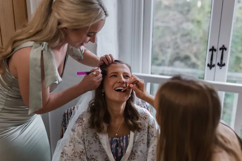 Bride smiling during fun bridal makeup moment with bridesmaids before ceremony