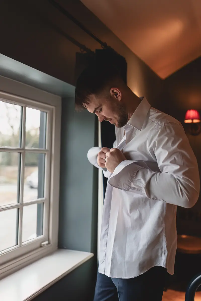 Groom fastening his shirt cuffs by the window during relaxed wedding morning preparations