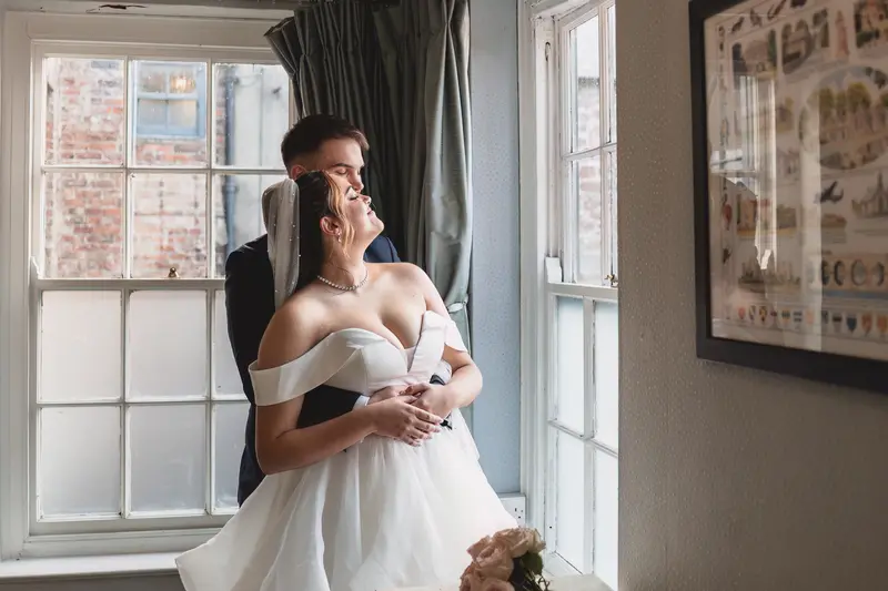 Bride and groom embracing beside a window during elegant natural light wedding portraits