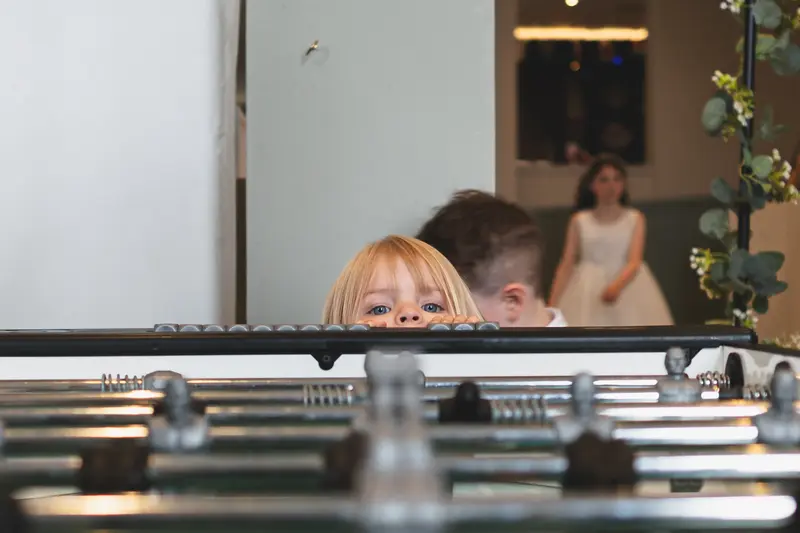 Curious child peeking over a table football game during a wedding reception, captured with natural documentary wedding photography