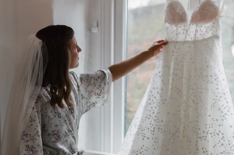 Bride admiring elegant wedding dress hanging by window before ceremony