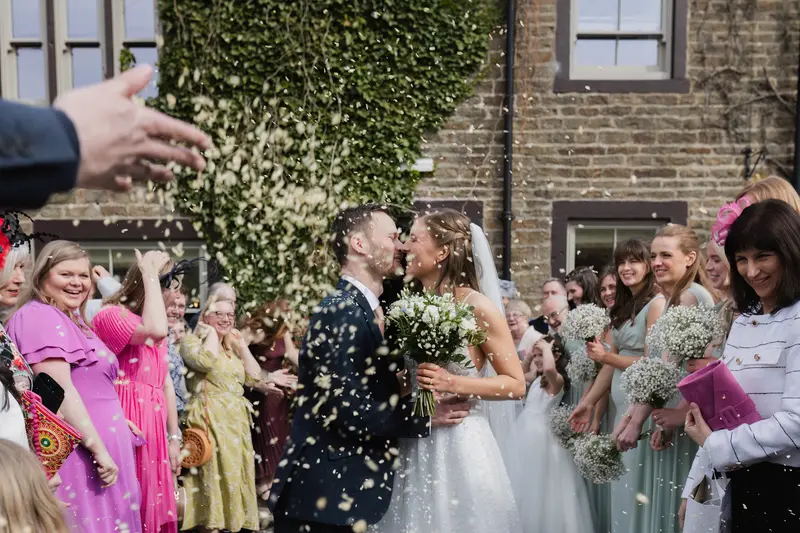 Documentary wedding confetti moment with newly married couple kissing outside Yorkshire wedding venue surrounded by cheering guests and bridesmaids