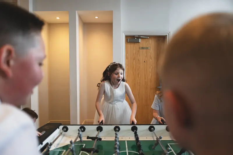 Excited flower girl playing table football during a wedding reception, captured in a natural documentary wedding photography style