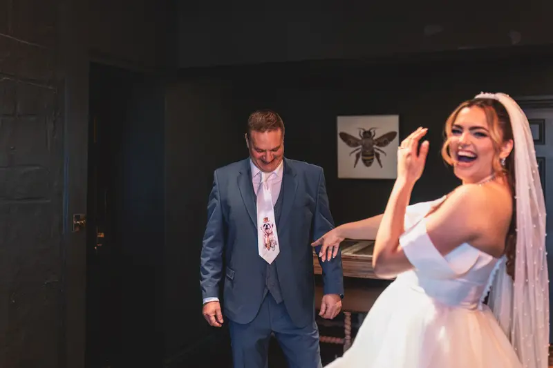 Bride laughing as her father reveals his personalised wedding tie during emotional wedding preparations